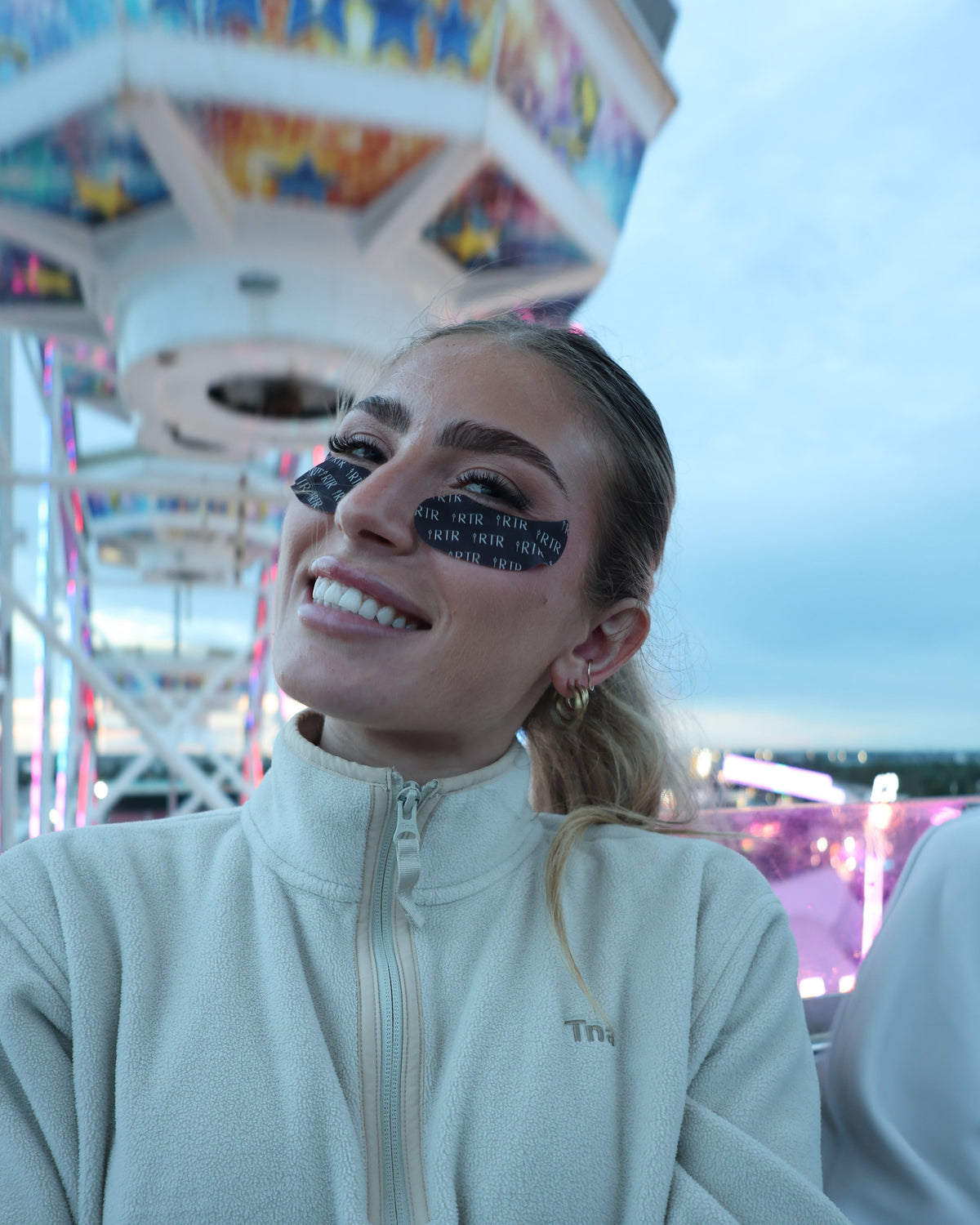 A woman smiling at an outdoor event with a carousel in the background, wearing the RTR depuffing eye patches