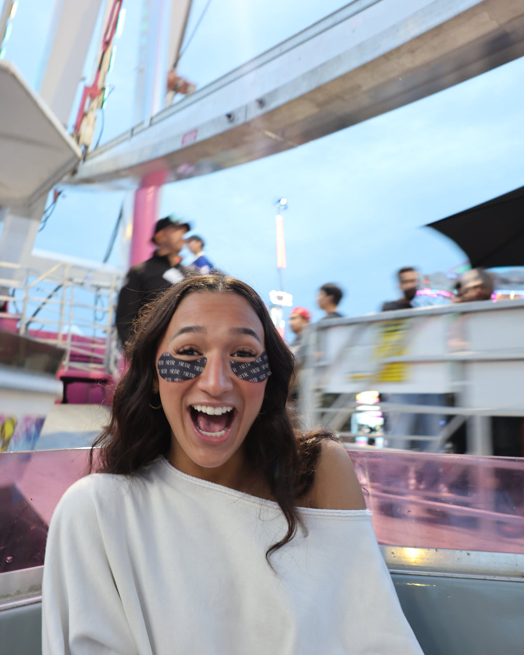 A woman with a wide smile and heart-shaped glittery eye stickers sits in a ride on a fairground or carnival ride with a Ferris wheel in the background.