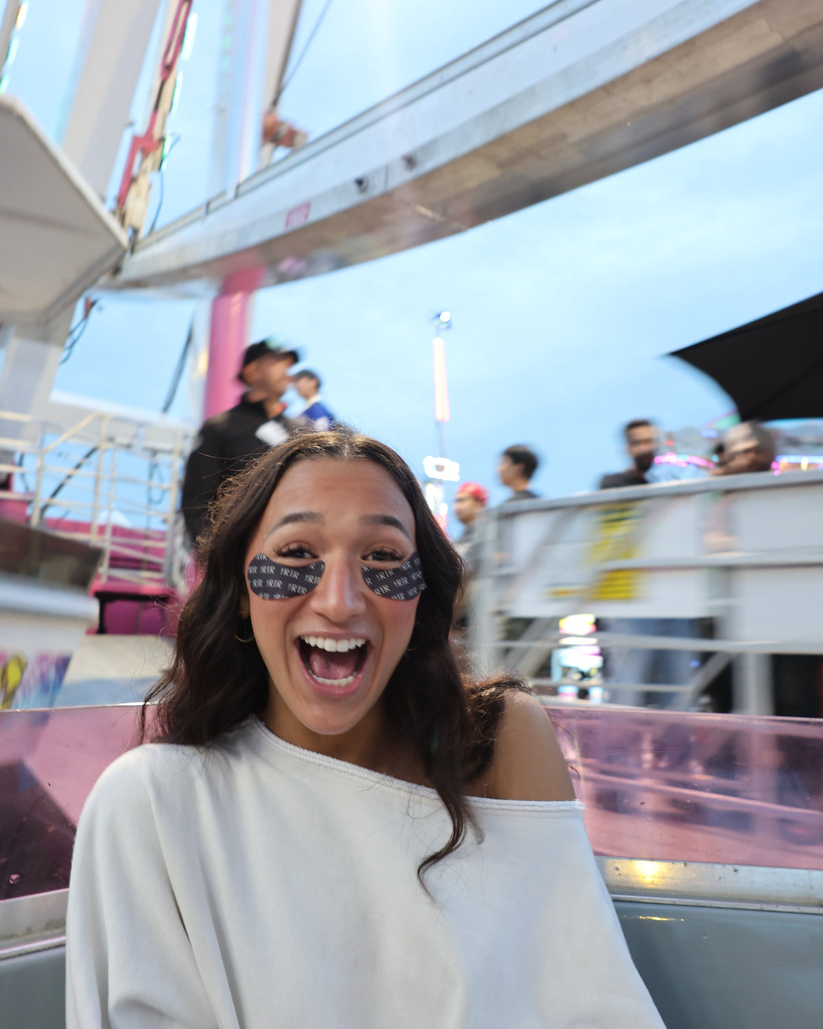 A woman with a wide smile and heart-shaped glittery eye stickers sits in a ride on a fairground or carnival ride with a Ferris wheel in the background.